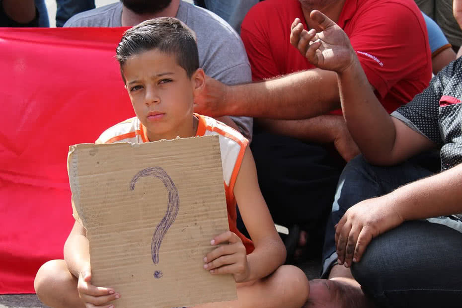 A Syrian boy and refugee holds up a homemade sign with a question mark.