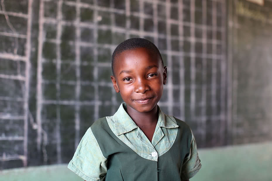 A schoolgirl stands in front of a blackboard in a classroom smiling in Zimbabwe.