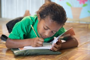 A school child lies on the classroom floor writing in school book